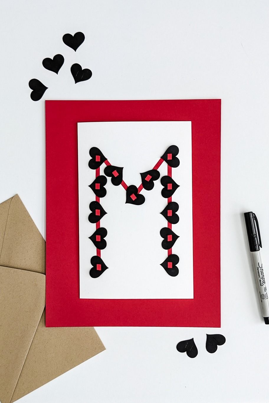 diy valentine with the initial m made of small black paper hearts strung along a red outline on a white card with red border