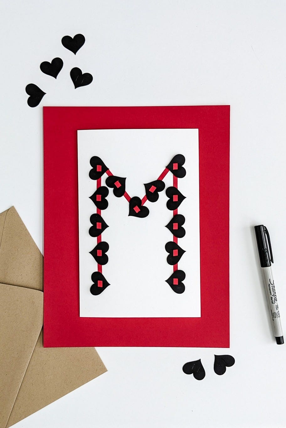 diy valentine with the initial m made of small black paper hearts strung along a red outline on a white card with red border