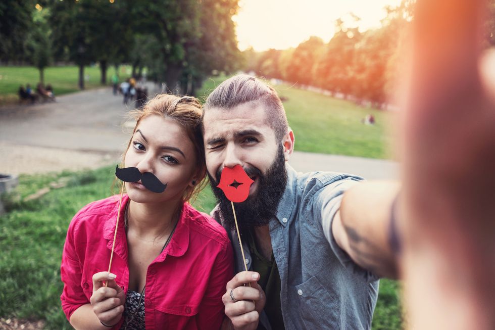cheerful teenage couple making funny selfies in the park, grimacing and holding paper mustaches and red lips
