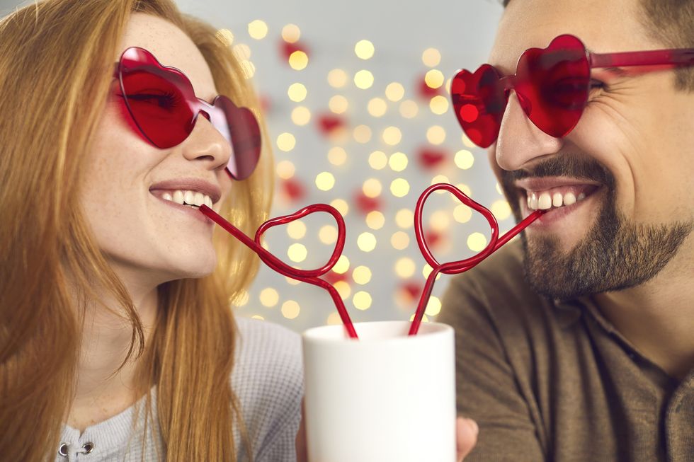 close up portrait happy young man and woman in love in pink sunglasses sipping drink from one cup through heart shaped straws, enjoying cute funny couple moment on a fun date on saint valentines day