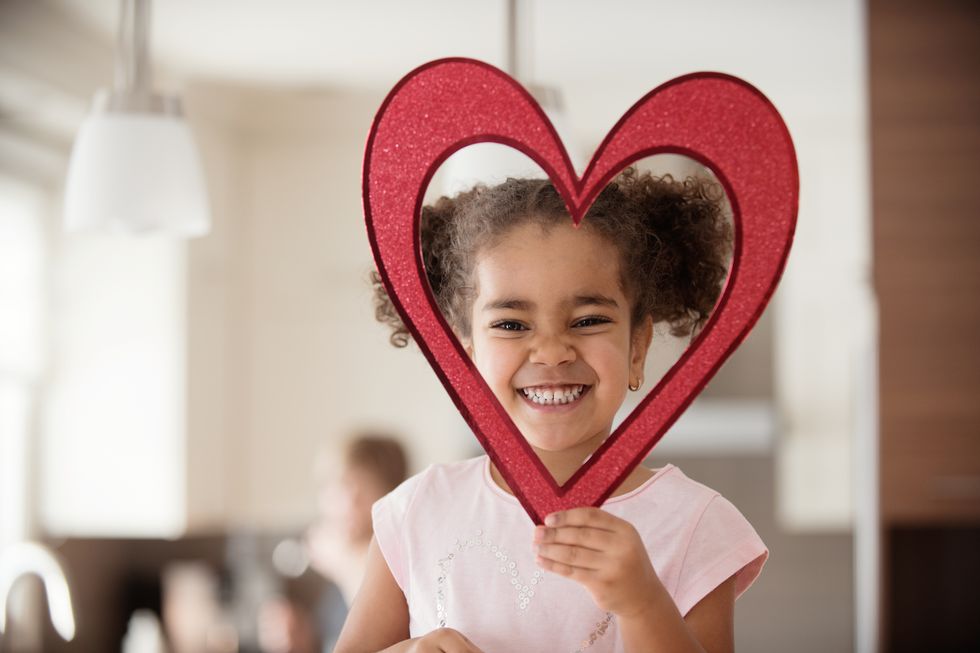 child holding a large glittery red paper heart with a cutout center that frames her face, she smiles adorably