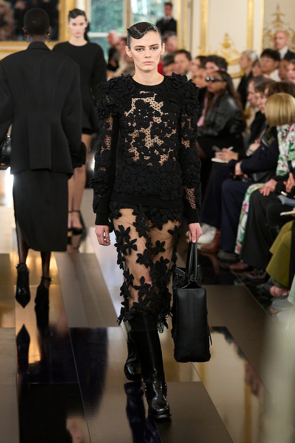 Fashion runway showcasing a model in a black floral outfit and kneehigh boots