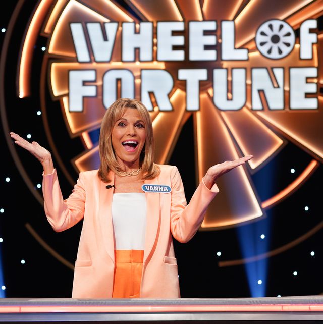 vanna white smiling with her arms in the air in front of the wheel of fortune logo