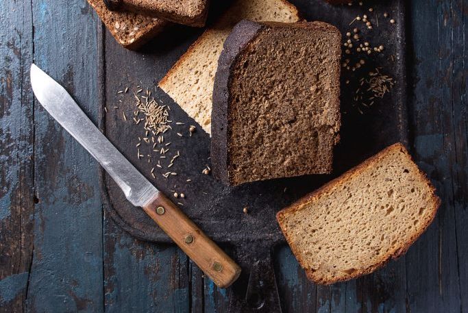 variety loaves of sliced homemade rye bread whole grain and seeds with knife on black cutting board over old dark wooden background top view, space healthy eating