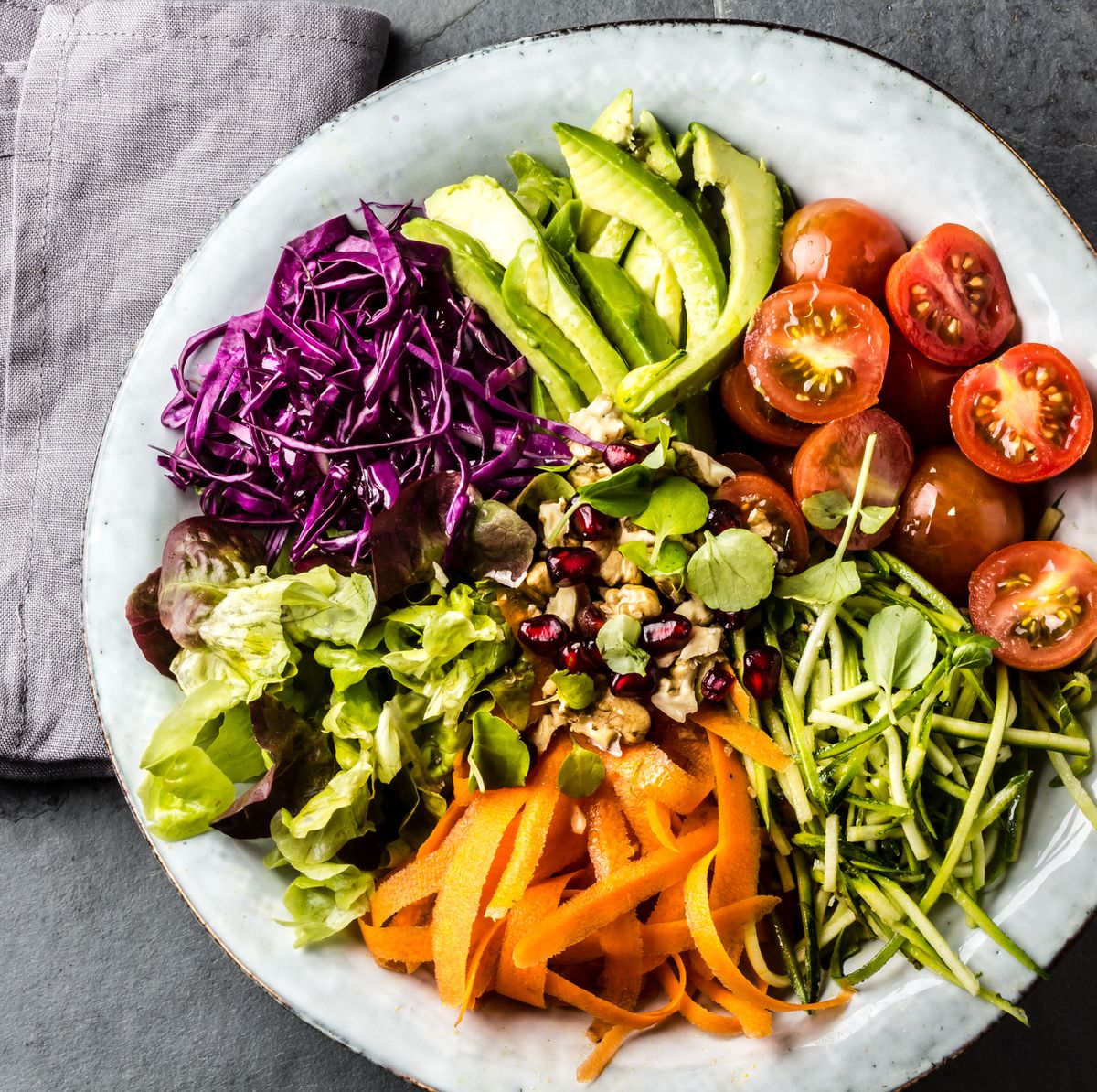 Vegan buddha bowl. Bowl with fresh raw vegetables - cabbage, carrot, zucchini, lettuce, watercress salad, tomatoes cherry and avocado, nuts and pomegranate