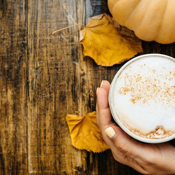 Vrouw vouwt handen om mok met pumpkin spice latte. Herfstbladeren en pompoen op houten achtergrond. 