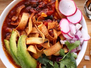 spicy chunky vegan tortilla soup is topped with red onions, avocado, tortilla chips, and cilantro in a white bowl against a wooden background