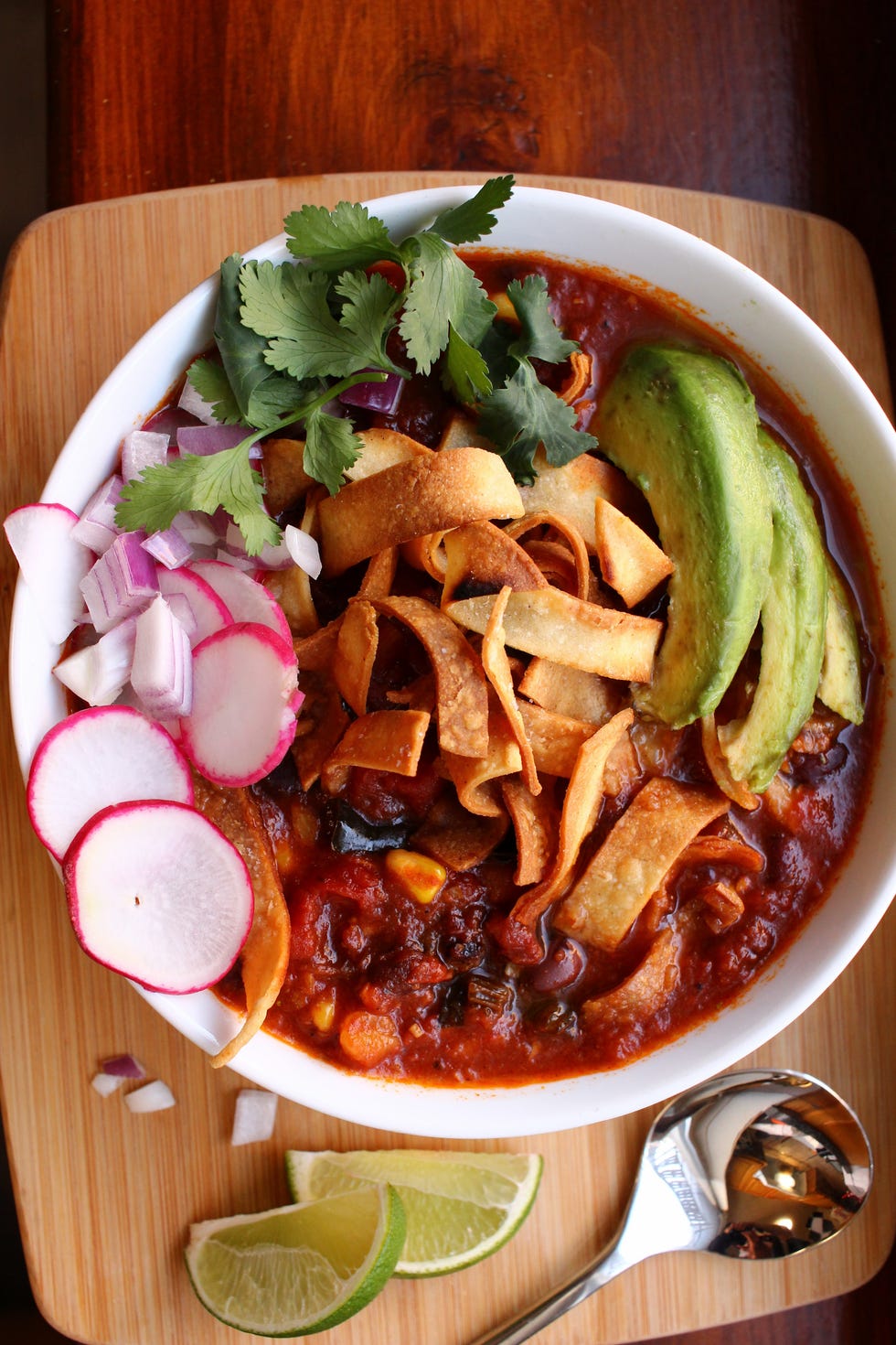 spicy chunky vegan tortilla soup is topped with red onions, avocado, tortilla chips, and cilantro in a white bowl against a wooden background