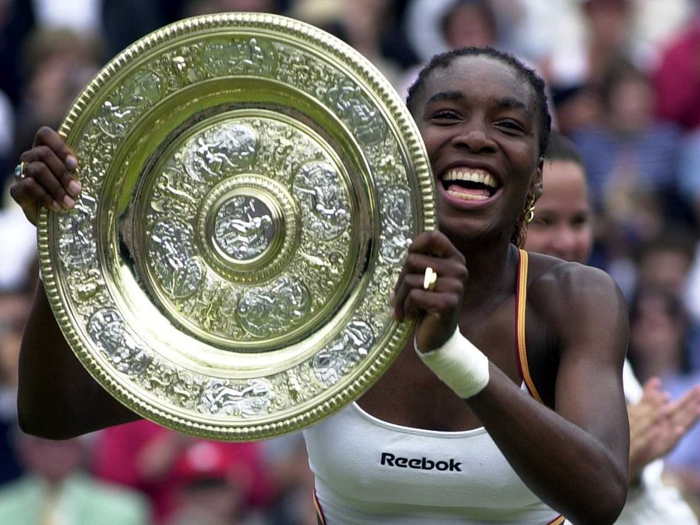 a woman smiles widely as she holds up a large platter trophy, she wears a white tennis outfit