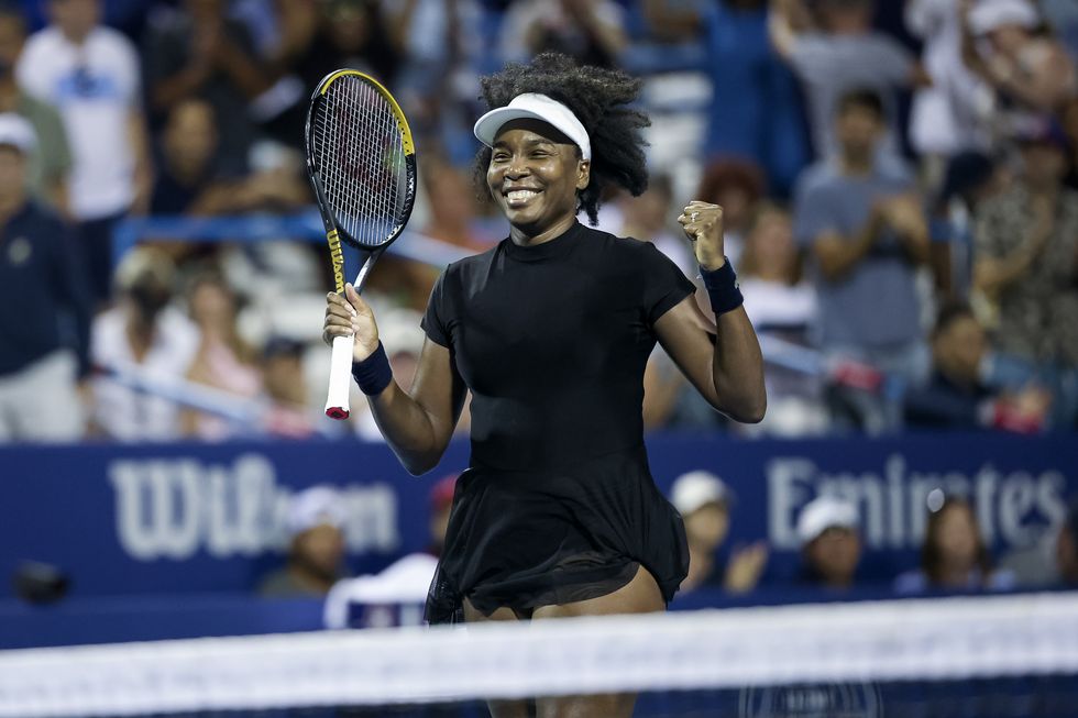 a woman smiles while holding a tennis racquet in one hand and pumping her other first, she wears all black and a white visor