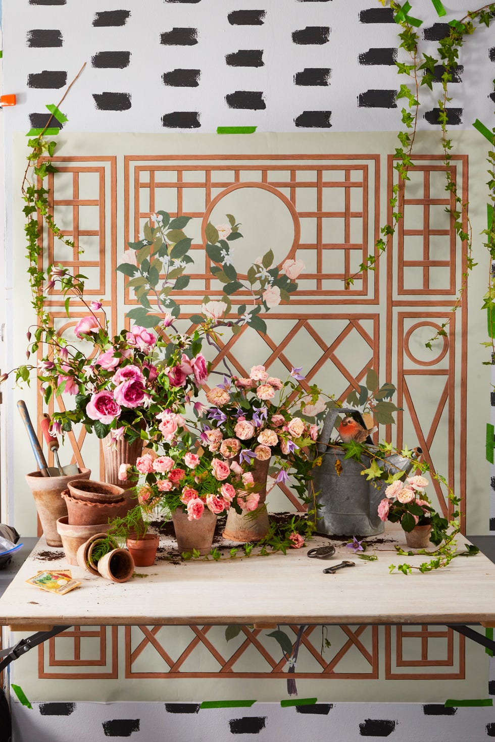 Floral arrangement with pots and gardening tools on a table against a painted backdrop