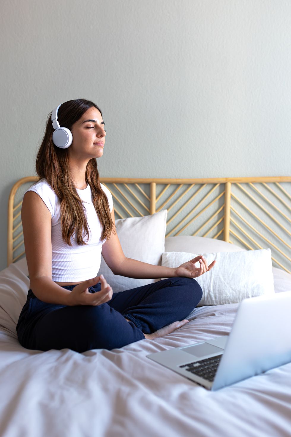 vertical portrait of young caucasian woman meditating at home sitting on the bed following online meditation, listening with headphones, using laptop
