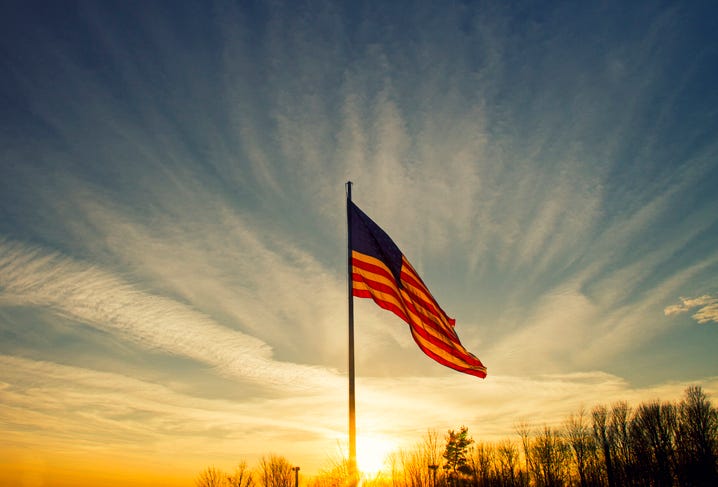 the sun sets behind a large flapping american flag under a blue sky