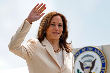 kamala harris waves and smilies at the camera, she wears a tan suit jacket over a white blouse and a golden chain necklace