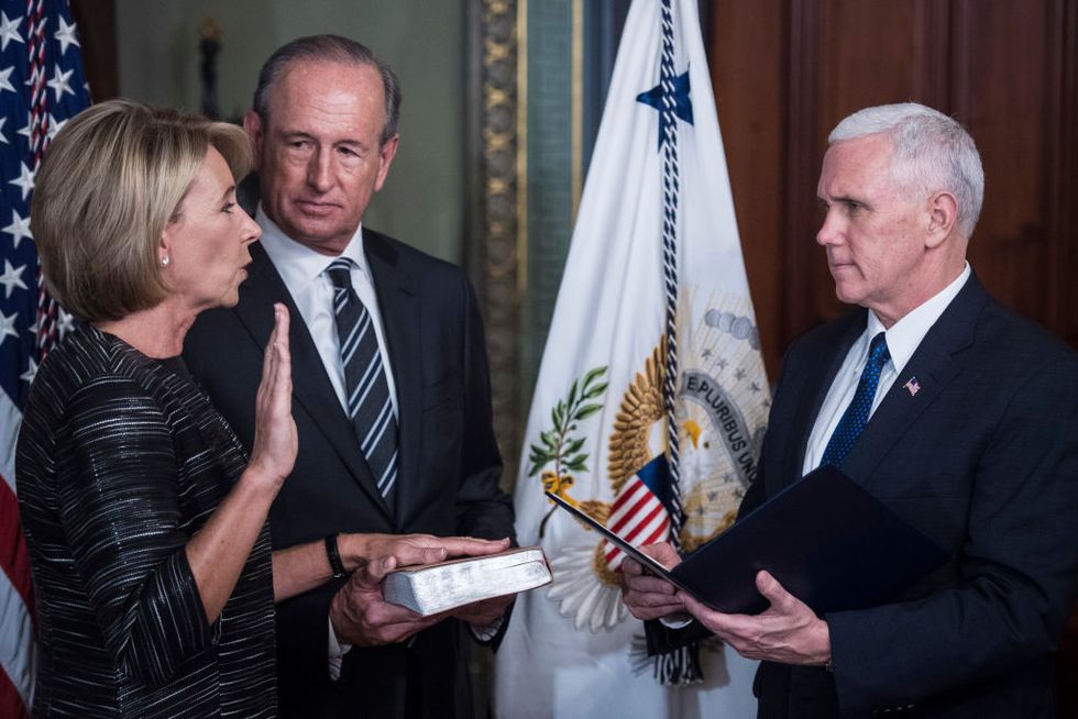 betsy devos stands with one hand raised and the other on a book held by husband dick devos, she looks at and speaks to mike pence who holds an open folder, behind them are two flags