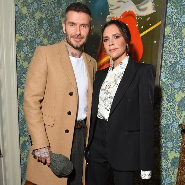 david beckham and victoria beckham stand and pose for a photo together, he wears a tan coat with a white shirt and dark pants, she wears a black suit with a white patterned shirt, they stand in front of a piece of artwork hanging on a wall with blue and green floral wallpaper