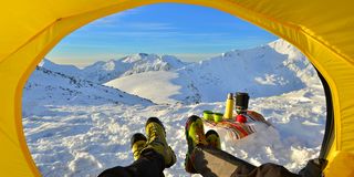 view from inside a yellow tent showing two peoples' legs with boots on, snowy mountains in distance