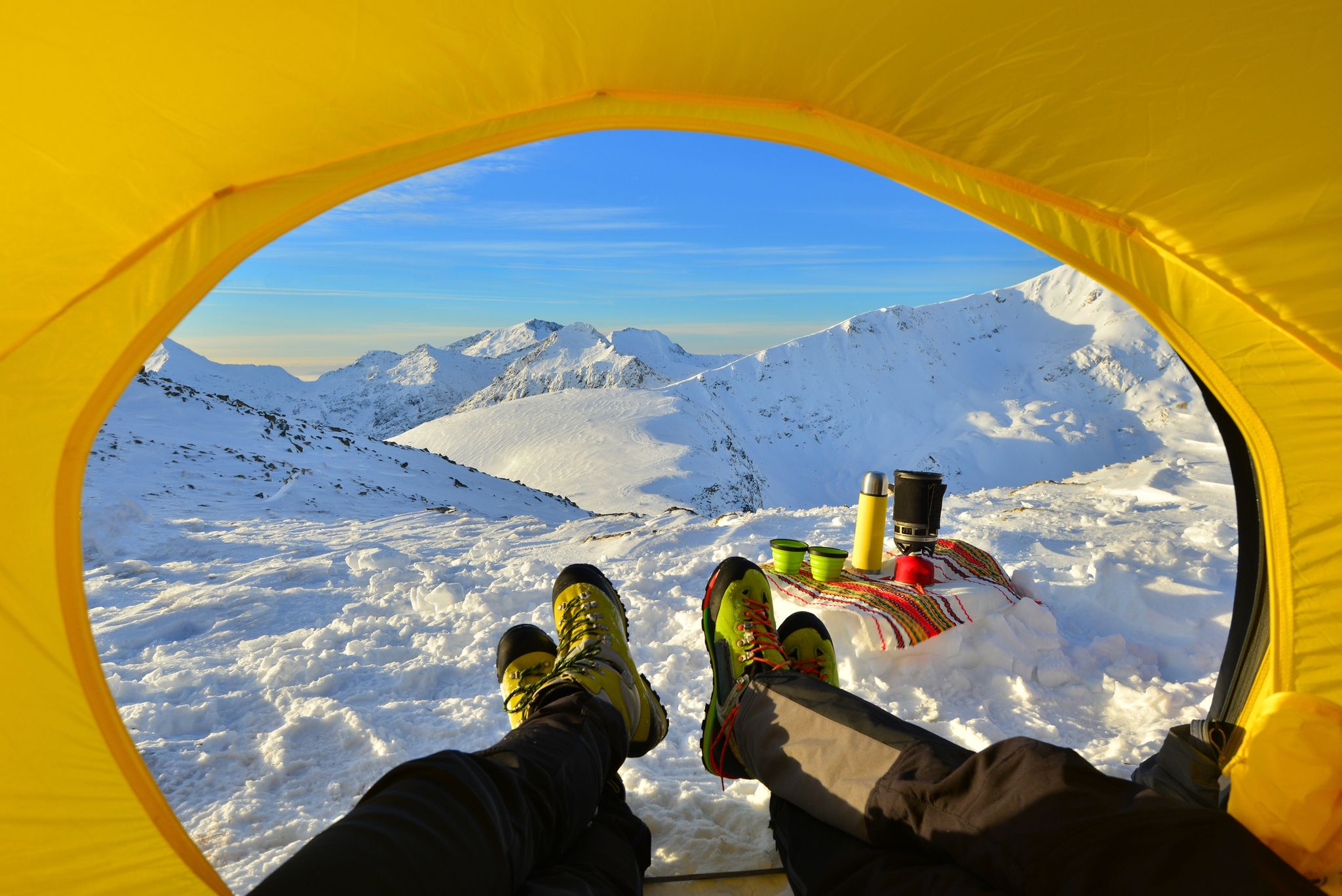 view from inside a yellow tent showing two peoples' legs with boots on, snowy mountains in distance
