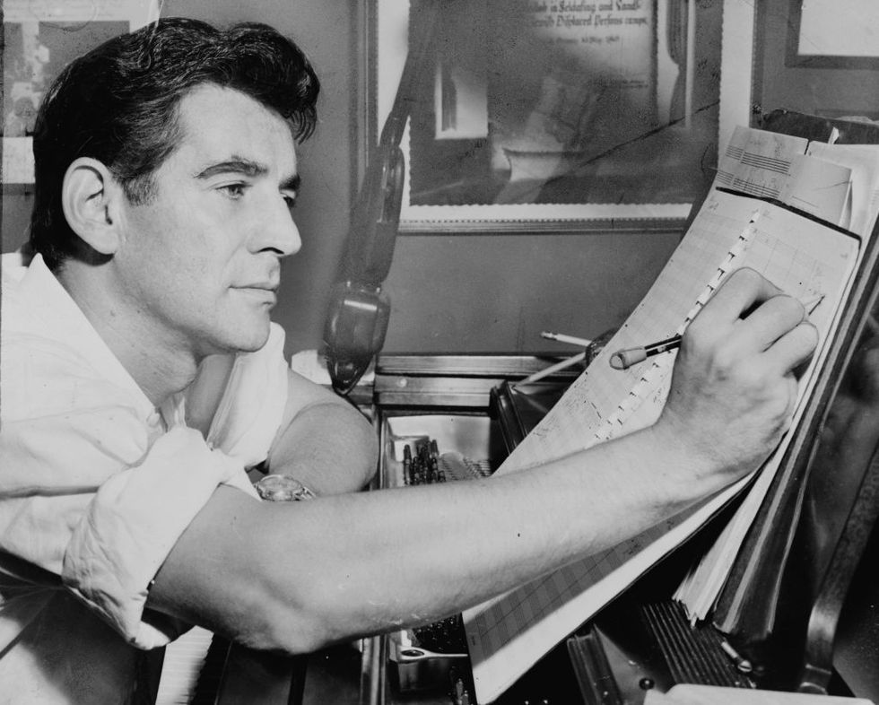 leonard bernstein sitting at a piano and making notations to a musical score in 1955