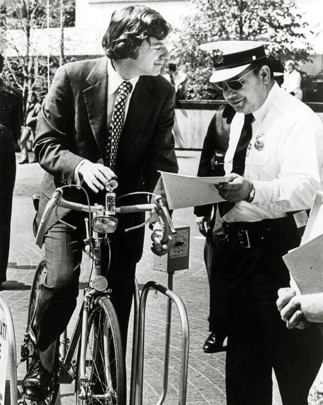 jerry springer stops while standing above his bicycle and talks to a police officer, springer is wearing a suit and a polka dot tie