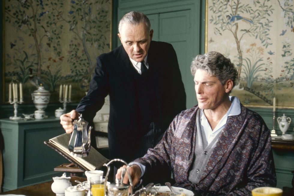 a man stands at a table and pours tea while another man sits at the table and watches, the table is full of dishes and food