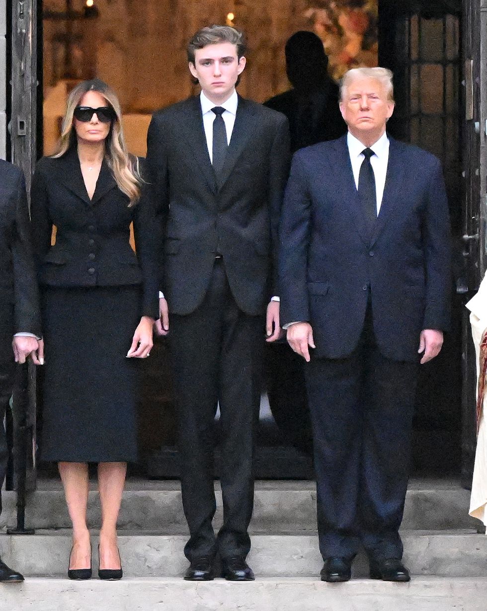 melania trump, barron trump, and donald trump stand on steps in front of an open set of doors, melania wears a dark skirt suit and sunglasses, barron and donald wear suits with ties and white collared shirts