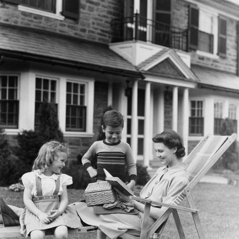 two children and mom sitting on a bench in front of a house, vintage black and white photo