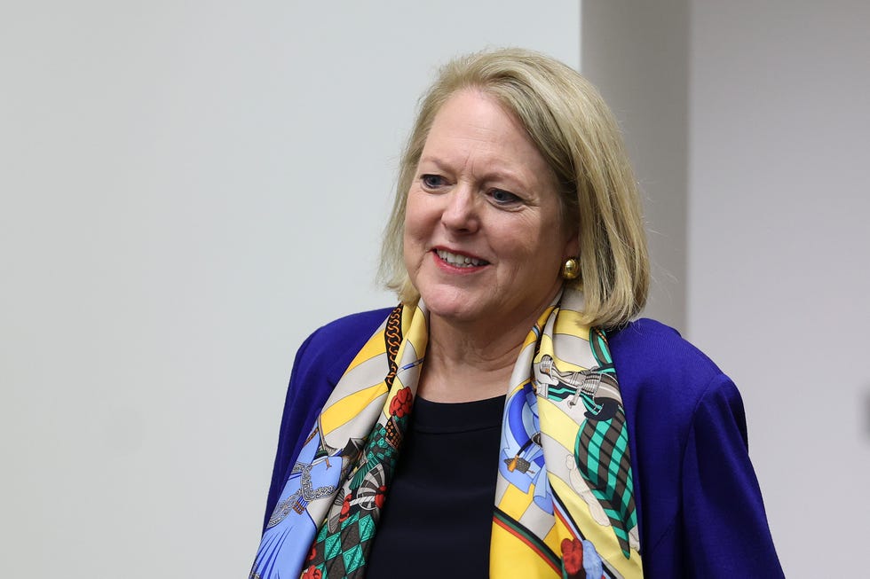 virginia thomas, wearing a blue pant coat and multicolored scarf, smiles and walks through a white hallway