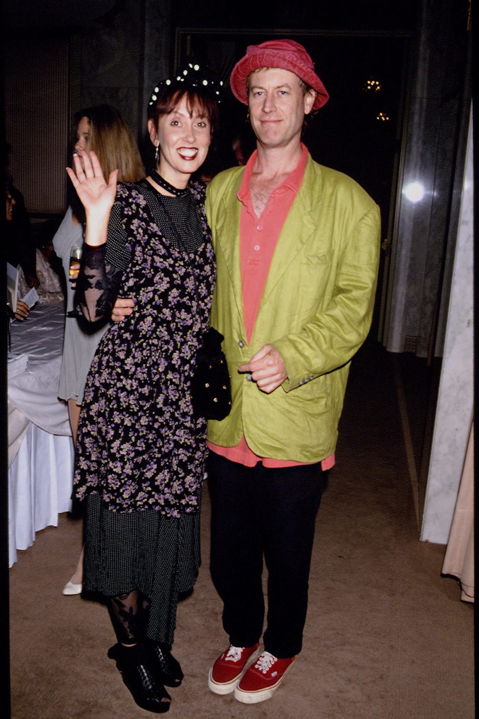 shelley duvall and dan gilroy stand together and smile at the camera, she waves one hand as they hold each other around the waist
