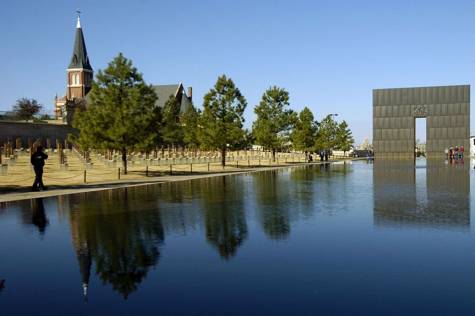 a reflection pool sits in the foreground, the background includes trees, a green lawn with identical statues, a church steeple, and a large square statue