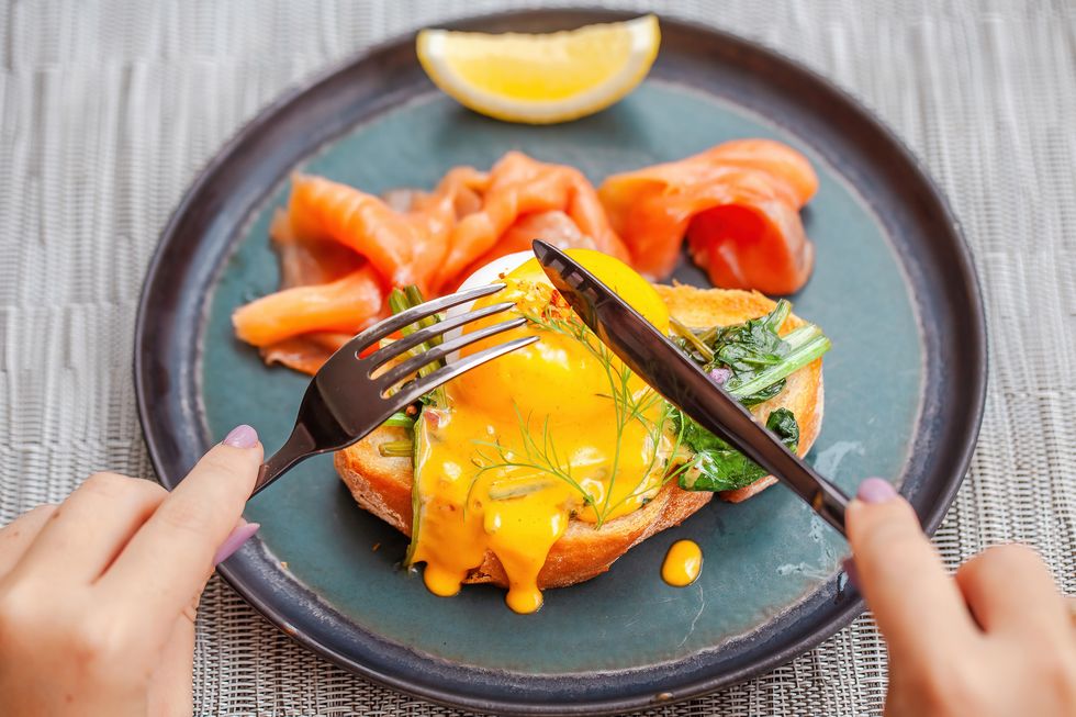 woman hands cut by fork and knife fresh eggs benedict with smoked salmon on plate in restaurant