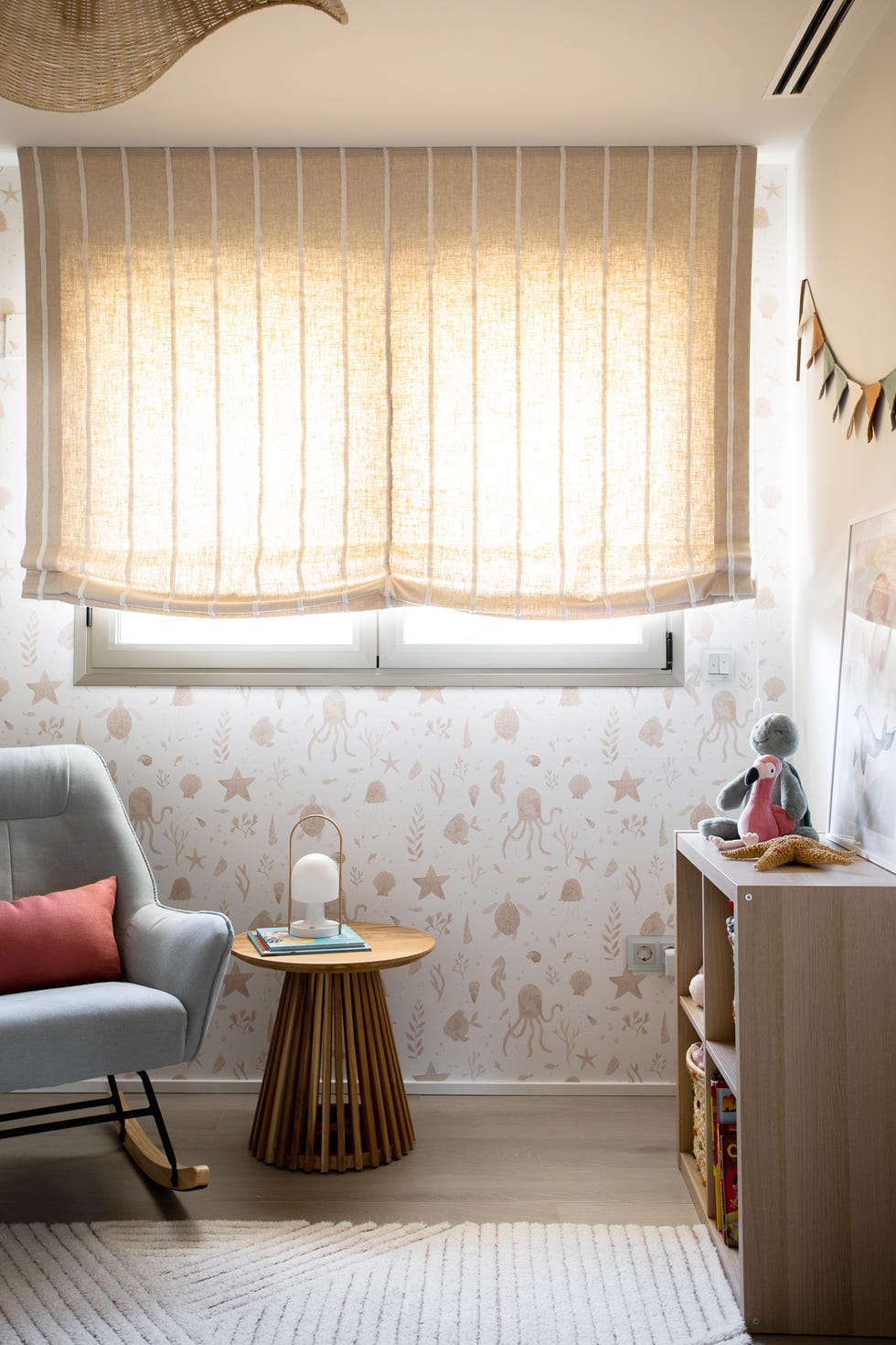 dormitorio infantil con mecedora, mesa auxiliar de madera redonda, librería baja de madera y papel pintado con motivos marinos
