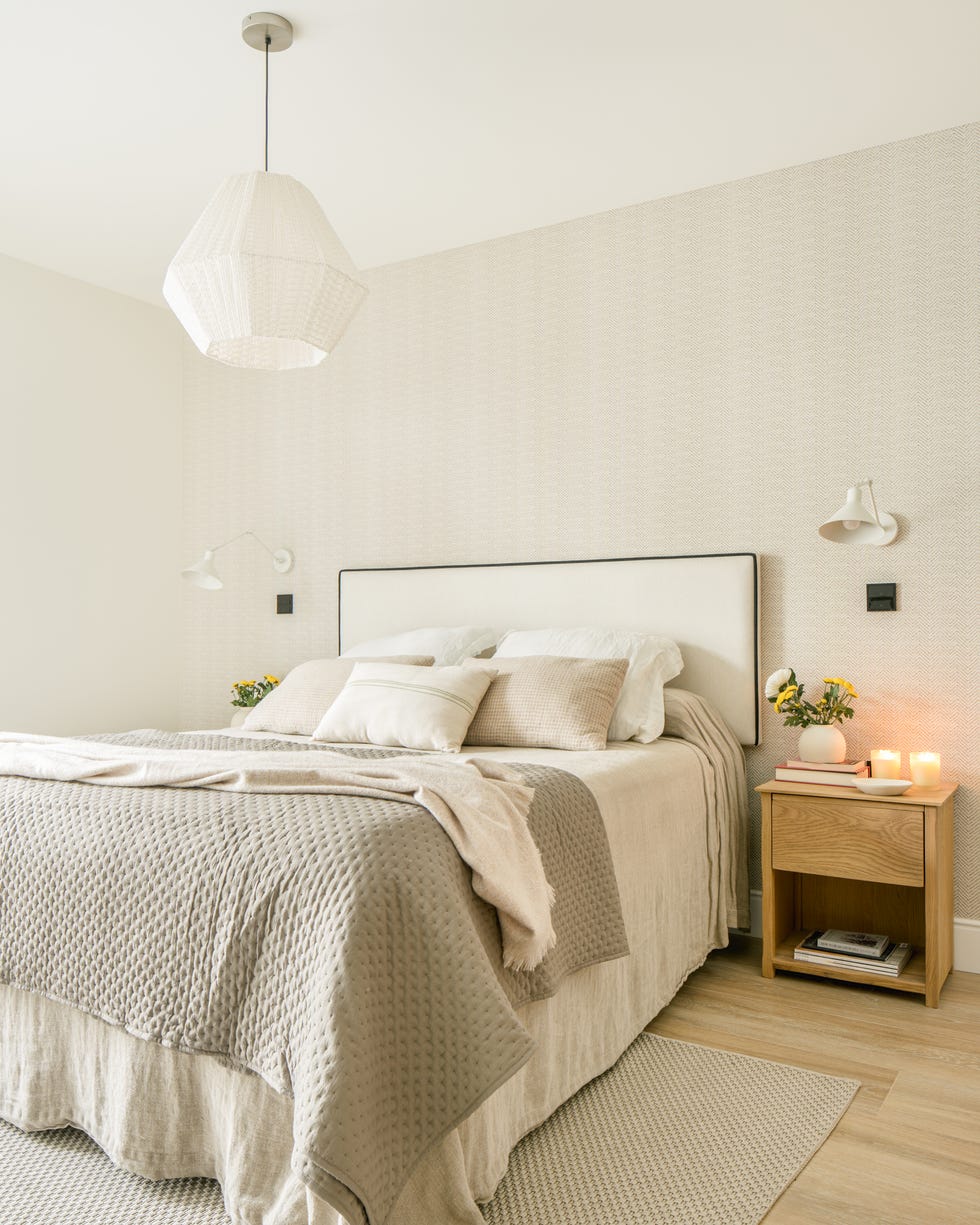 dormitorio con papel pintado efecto tejido, cabecero de cama tapizado en beige con ribeteado negro y mesitas de noche de madera con apliques de pared