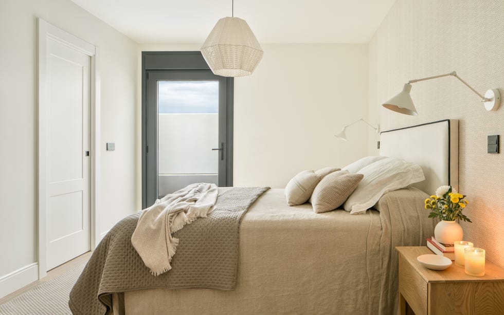 dormitorio con papel pintado efecto tejido, cabecero de cama tapizado en beige con ribeteado negro y mesitas de noche de madera con apliques de pared