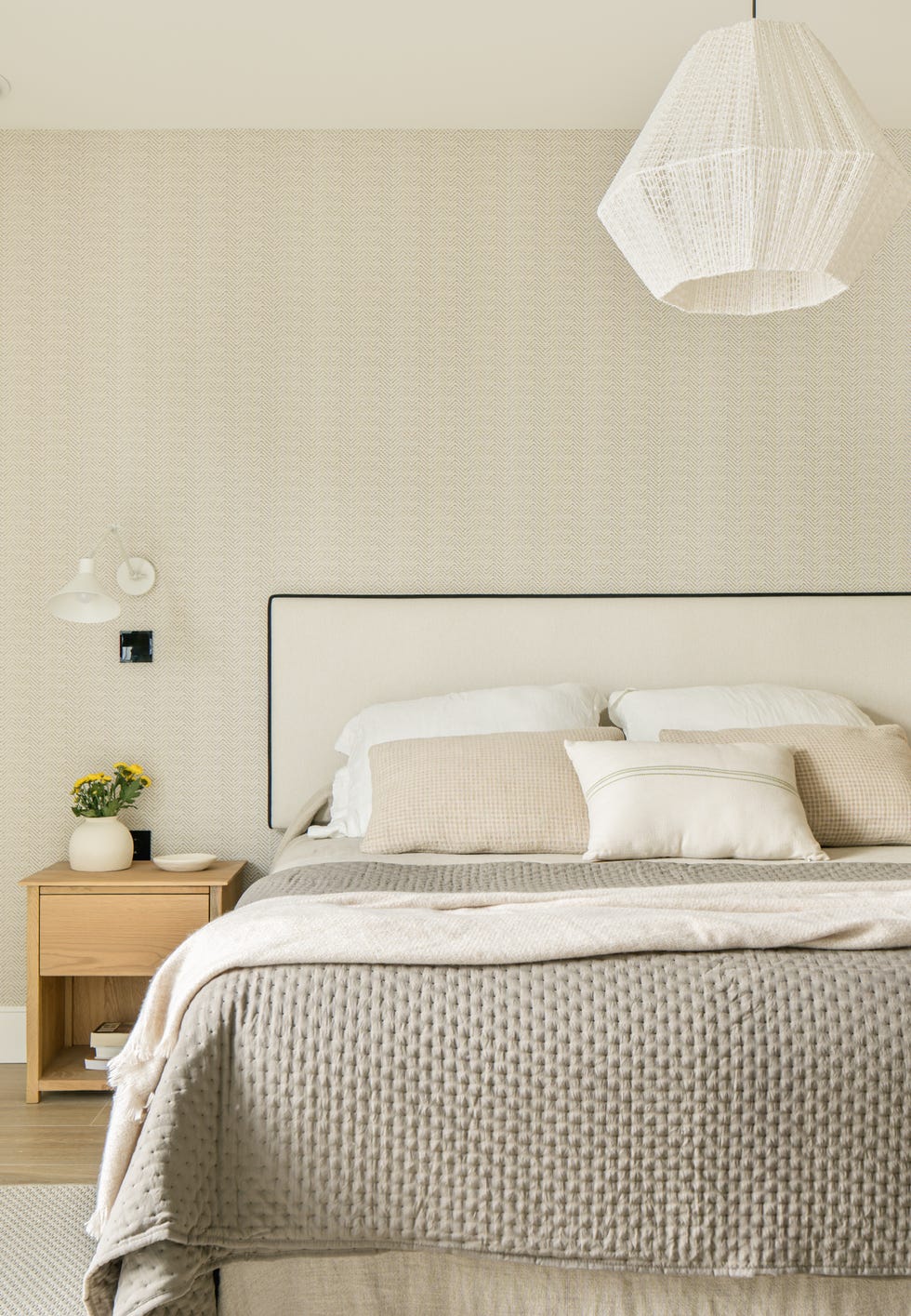 dormitorio con papel pintado efecto tejido, cabecero de cama tapizado en beige con ribeteado negro y mesitas de noche de madera con apliques de pared