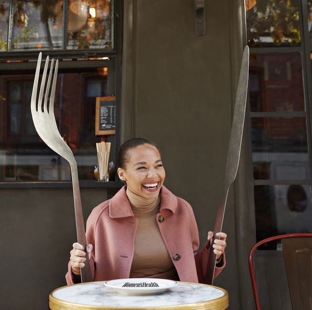 happy young woman holding large fork and table knife while sitting at sidewalk cafe