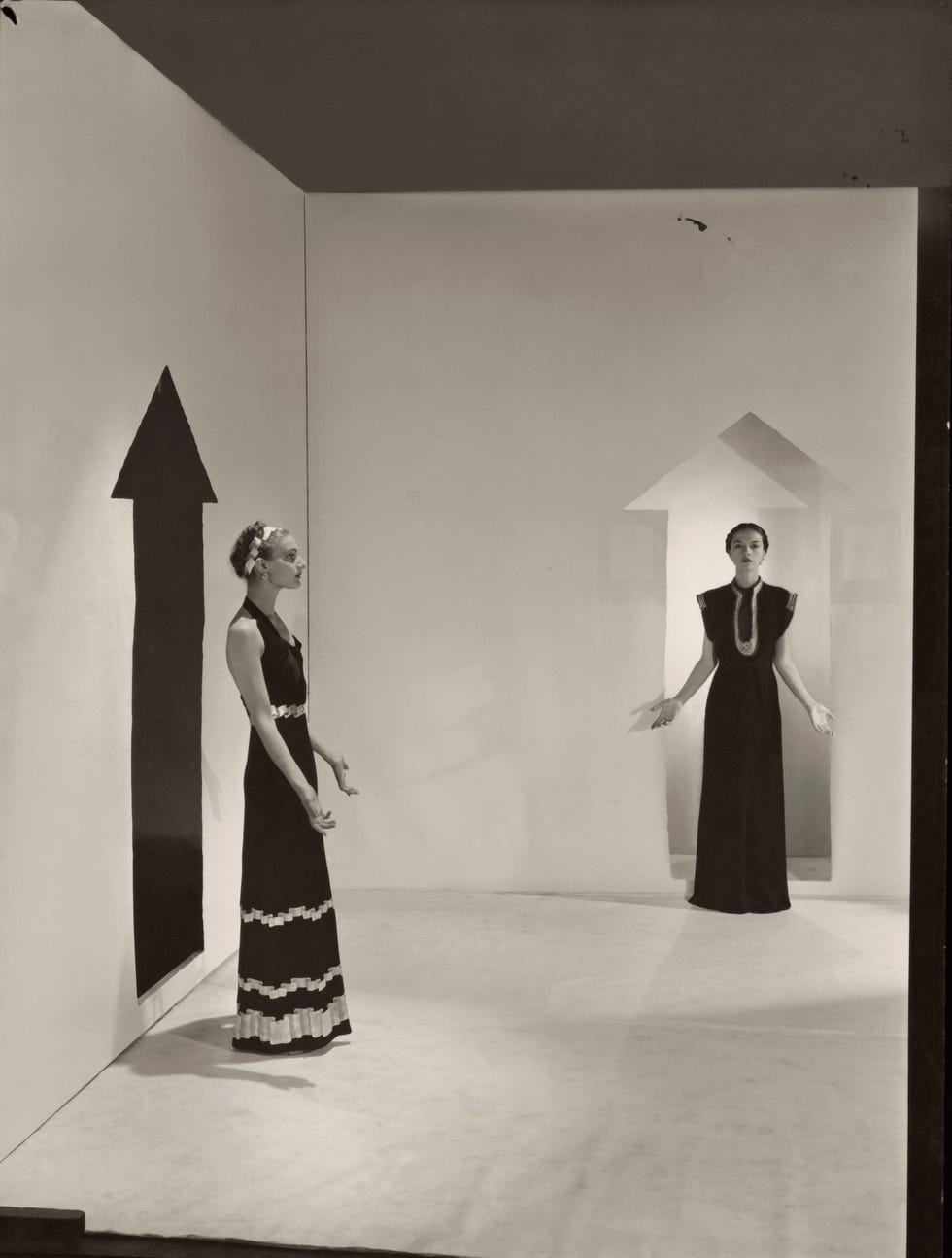 two models, standing in a white room with arrows painted on walls and wearing dresses by schiaparelli; left in a dark crepe with bands of gold braids down the front, over satin pajamas; right, in a black satin with bands of rippled gold ribbons. (photo by cecil beaton/condé nast via getty images)