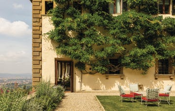 terrace of a florentine hotel with lush greenery wrapped around the walls, lawn chairs with bright red cushions, and views over the tuscan hills