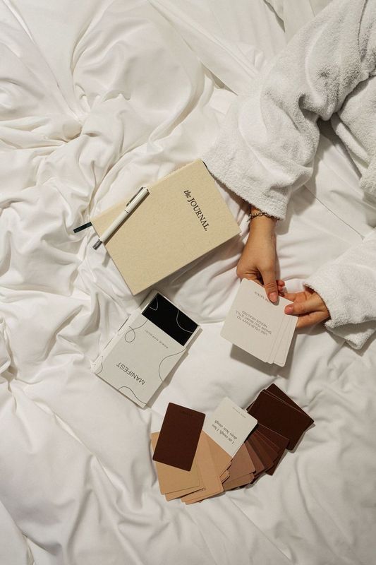 a person in a bathrobe holding a box of manifesting cards in a cosy bed with a crisp white duvet