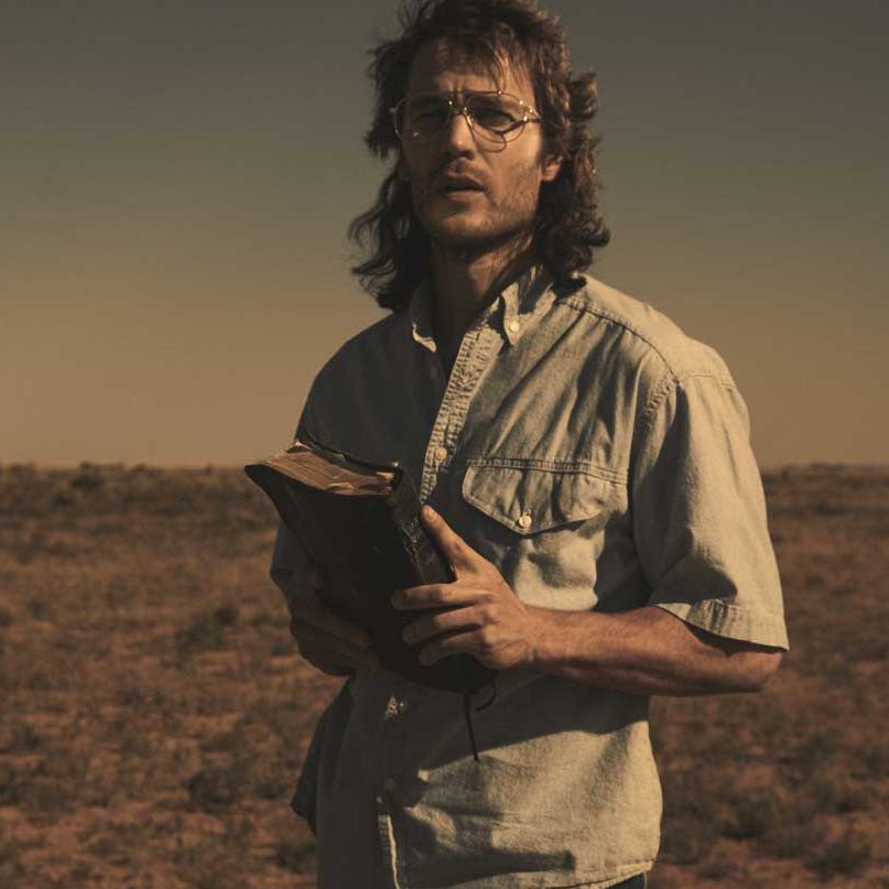 taylor kitsch as david koresh in the series, 'waco' he is standing in a deserted landscape with what appears to be a bible in his hand