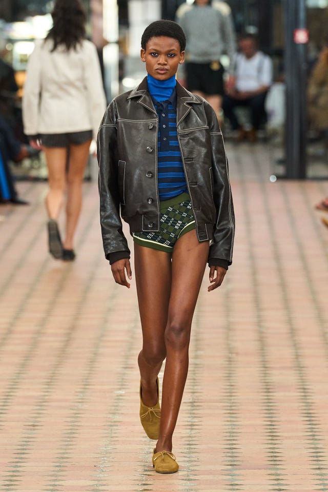 Fashion model walking down a runway wearing a brown leather jacket and patterned shorts