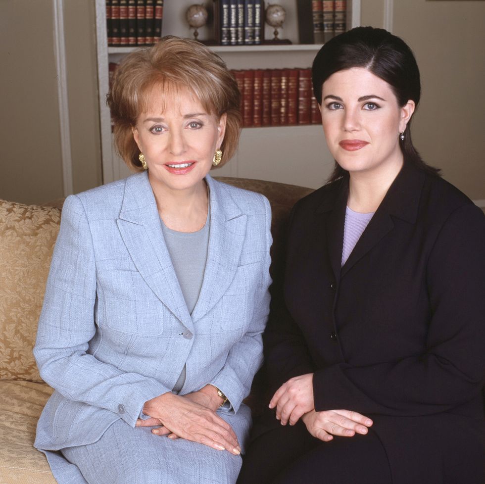 barbara walters and monica lewinsky sit on a sofa and smile at the camera, walters is wearing a blue suit with gold hoop earrings, lewinsky is wearing a black suit with a purple shirt and pearl earrings, behind them is a built in bookshelf
