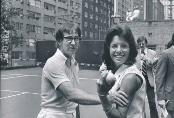 bobby riggs squeezes billie jean kings flexed bicep as she holds a tennis ball in her hand, both look right of the camera and stand on a tennis court, riggs is caught talking and king smiles