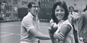 bobby riggs squeezes billie jean kings flexed bicep as she holds a tennis ball in her hand, both look right of the camera and stand on a tennis court, riggs is caught talking and king smiles