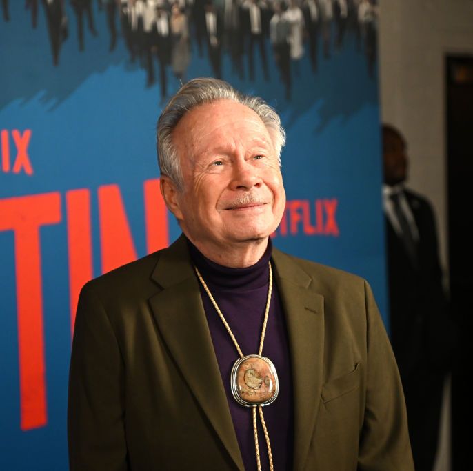 walter naegle smiles and looks past the camera, he wears a green suit jacket, black turtleneck and large pendant necklace