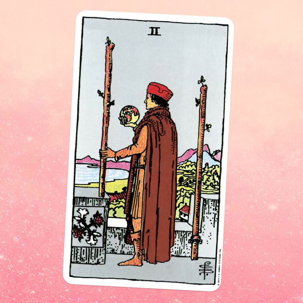 the tarot card the two of wands, showing a person in a tunic, cape, and robe, holding a glass ball they're standing between two wooden staffs, looking out at a view of a mountain and a body of water