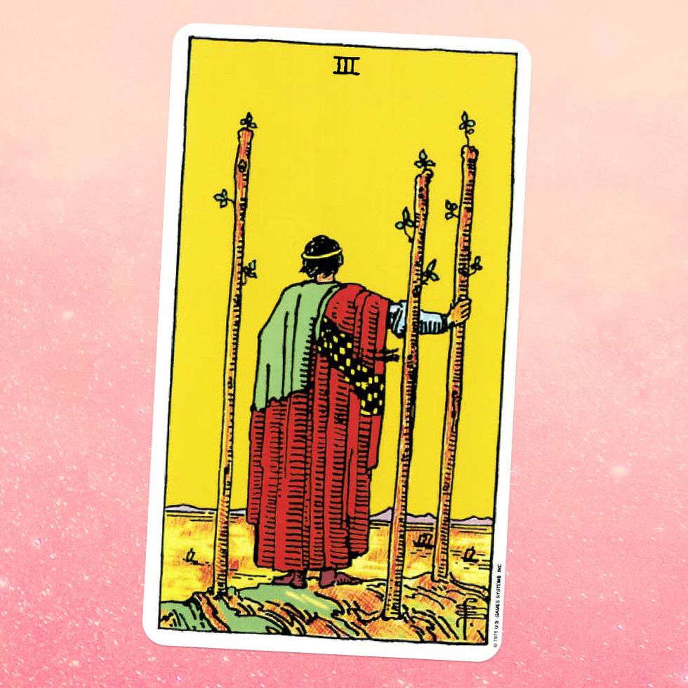 the tarot card the three of wands, showing a view of the back of person in a robe who is holding a wooden staff and looking out on a landscape, with two more wooden staffs growing out of the ground behind them