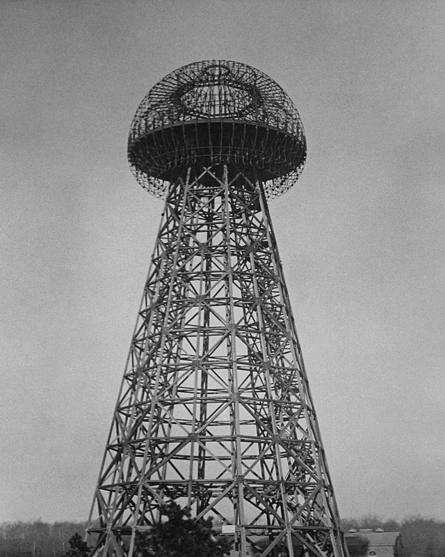 a large metal tower with a bulbous top stands outside, a building and trees are in the background