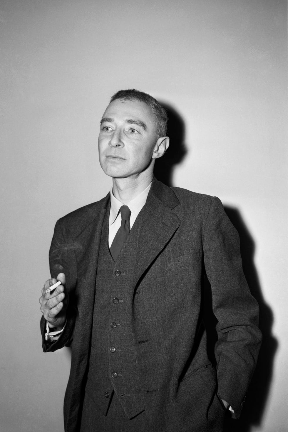 j robert oppenheimer holds a cigarette in one hand and has his other hand in another pocket, he wears a three piece suit