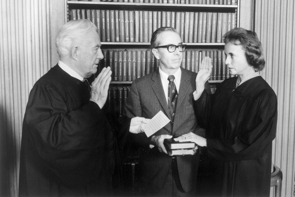 warren burger and sandra day o'connor stand facing each other each with their right hand raised, a man stands in the middle holding a stack of books that o'connor rests her left hand on